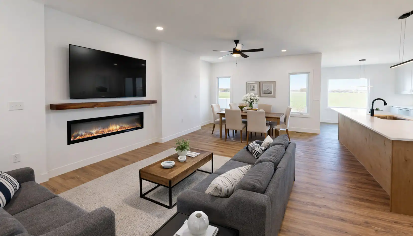 Modern open-concept living room with a gray sofa, wall-mounted TV, electric fireplace, dining area, and kitchen with white cabinets and wood flooring—perfect for a Leo seeking a stylish space to relax or entertain guests