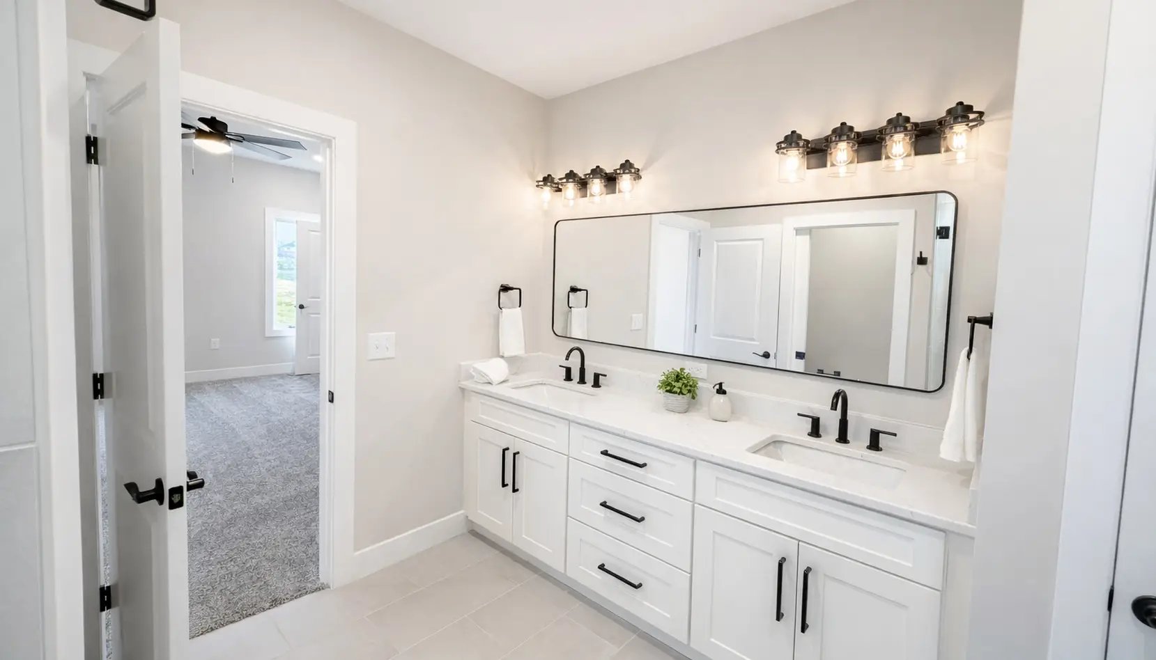 Modern bathroom with double sinks, a large mirror, black fixtures, white cabinets, and bright lights. The Asher-inspired design features a door leading to a carpeted bedroom, and the space is clean and neatly organized with minimal decor