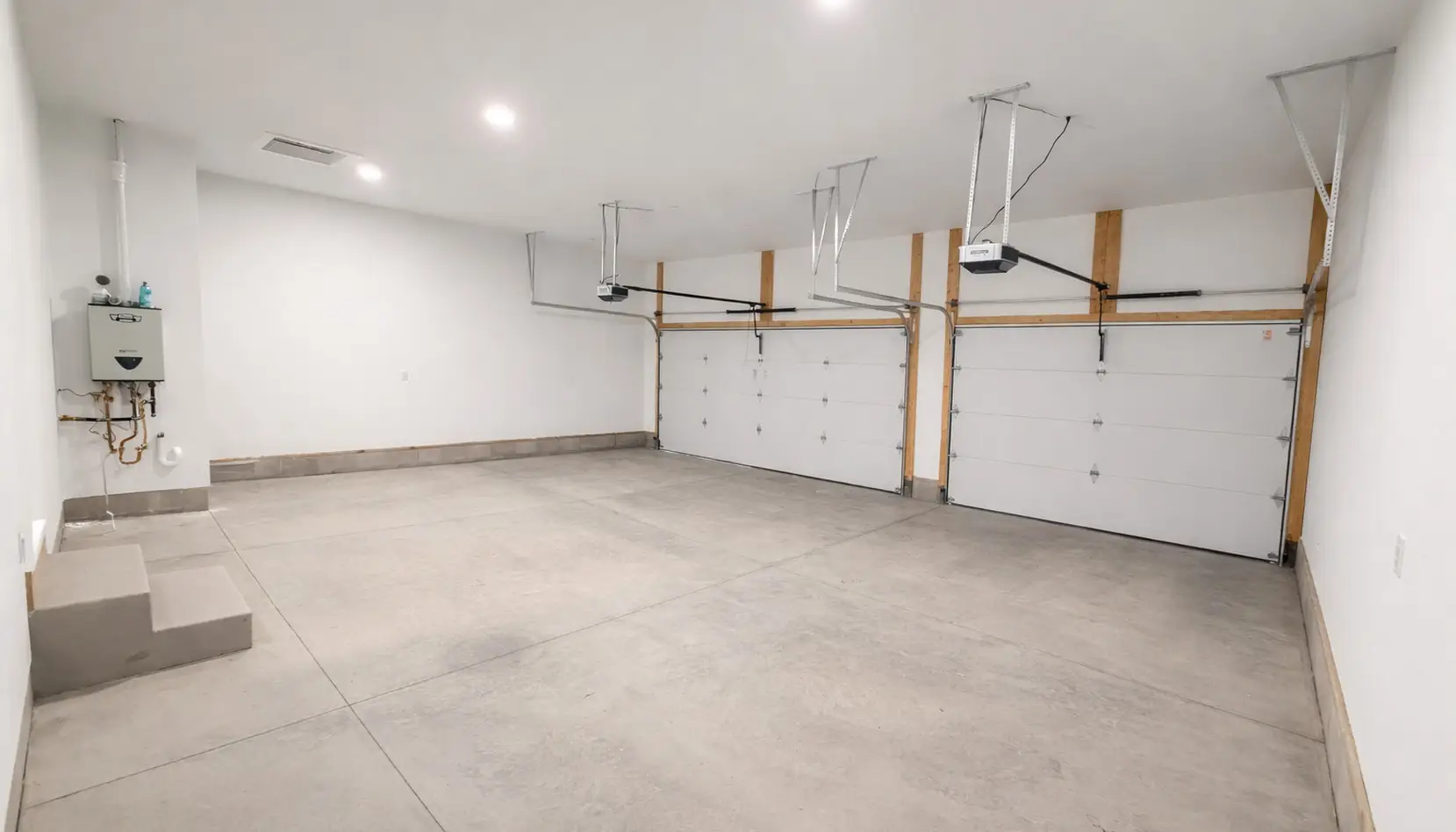 Spacious, clean garage with two closed white garage doors, concrete floors, and bright overhead lighting. Exposed piping and a water heater are mounted on the left wall, while a small set of steps leads to the door—making this Asher-inspired space both functional and inviting