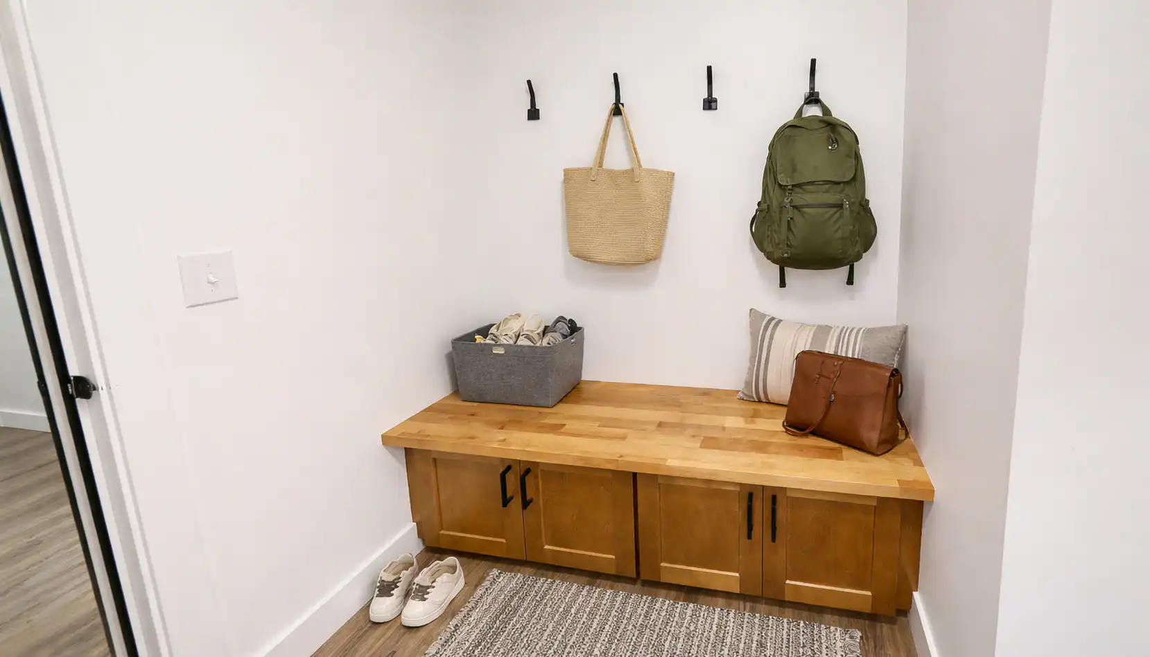 A small mudroom with a wooden bench, gray basket, pillows, shoes, a purse, and a green Leo backpack alongside a straw tote hanging on black hooks. The floor features light wood paired with a gray textured rug