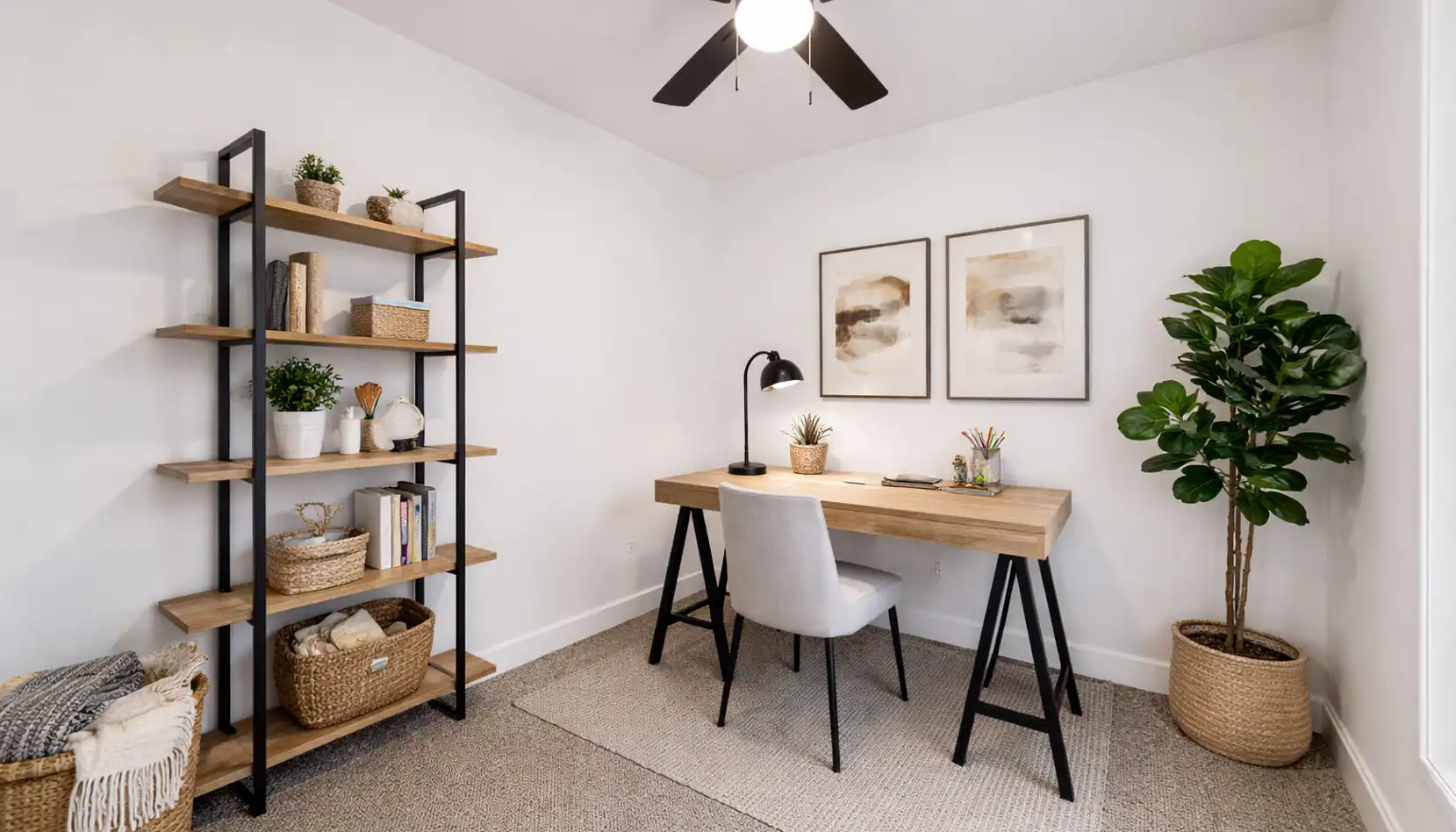 Minimalist home office with a wooden desk, beige chair, black lamp, wall art, bookshelf with baskets and decor, potted plant, and neutral tones. A ceiling fan spins above this cozy Leo-inspired space—organized, well-lit, and filled with subtle touches of personality