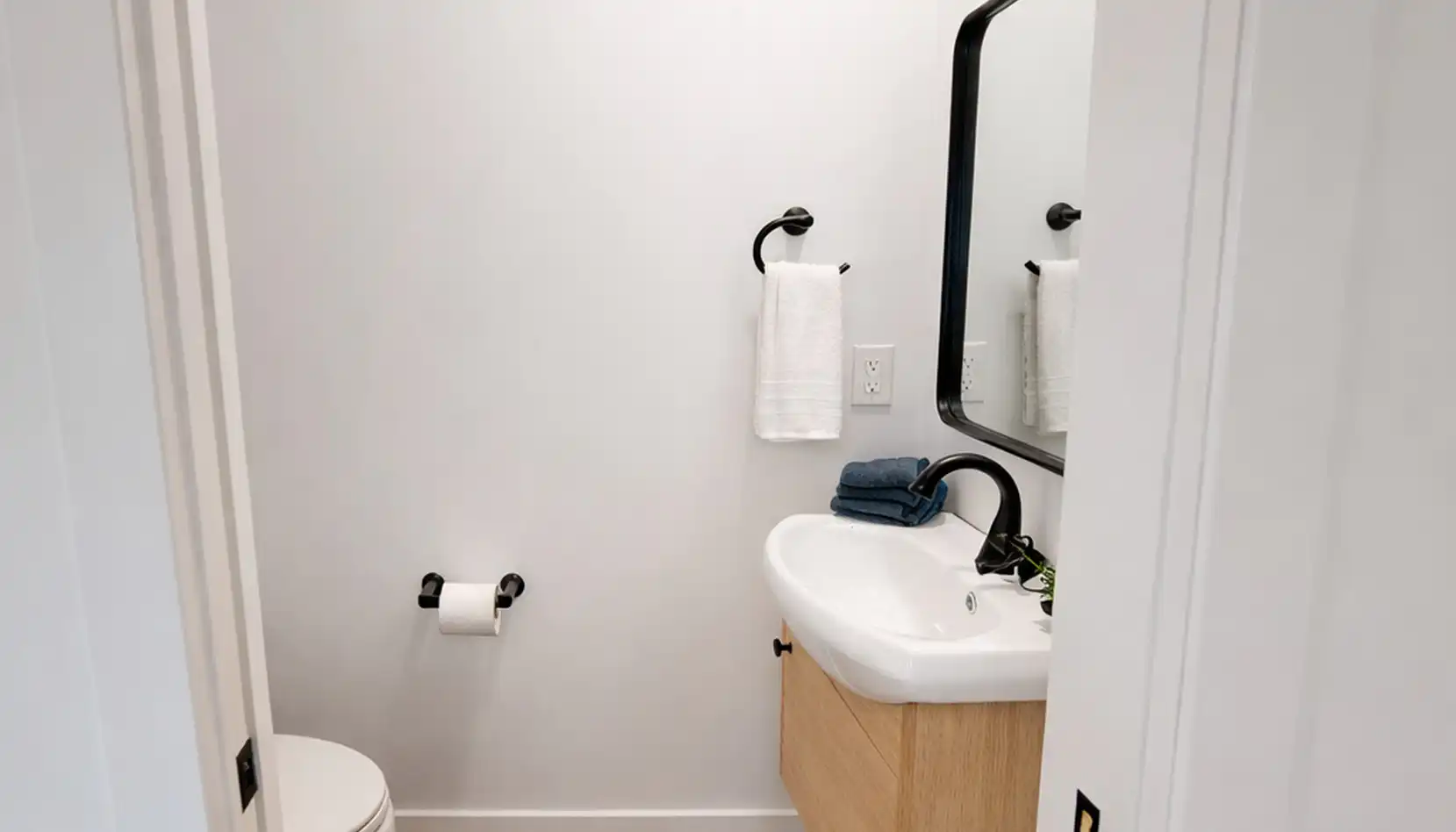 A modern bathroom with a white toilet, a towel ring holding a white towel, a wall-mounted sink with a black faucet, a stack of towels, and a rectangular mirror above the sink creates an inviting space reminiscent of Leo’s refined sense of style