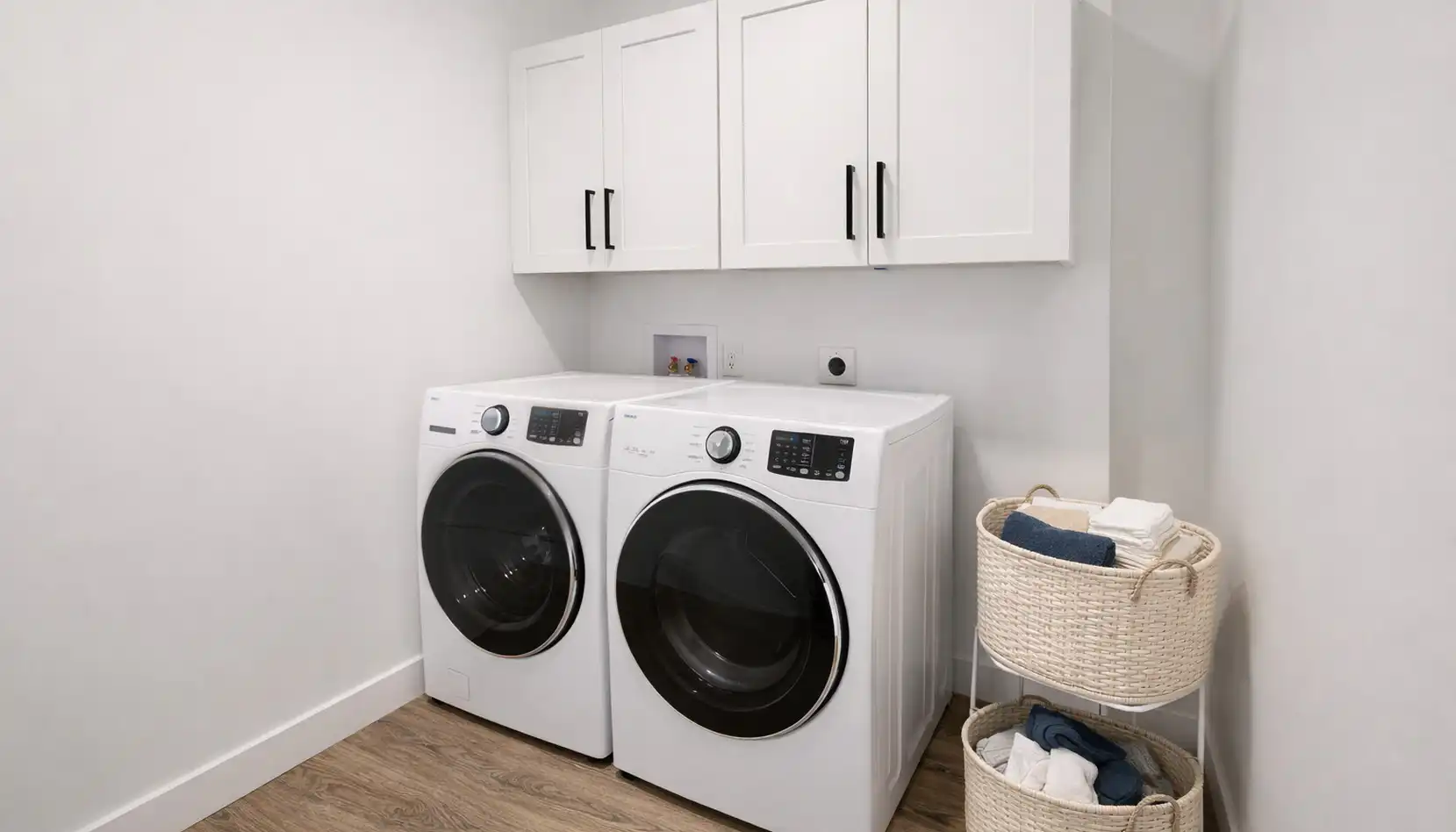 Modern laundry room with a white front-loading washer and dryer, white cabinets above, and a wicker basket stand holding folded laundry on the right. The floor is wood, and the walls are plain white, creating a clean and organized space that even Leo would appreciate for its simplicity and functionality