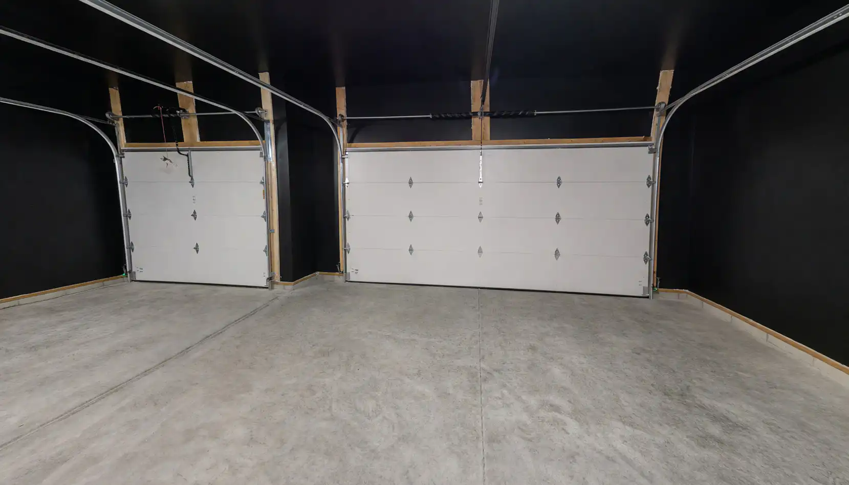 A clean, empty garage with two closed white overhead doors, black walls, exposed wooden beams, and a smooth concrete floor awaits—its modern simplicity accentuated by a bold Leo energy that brings warmth to the space
