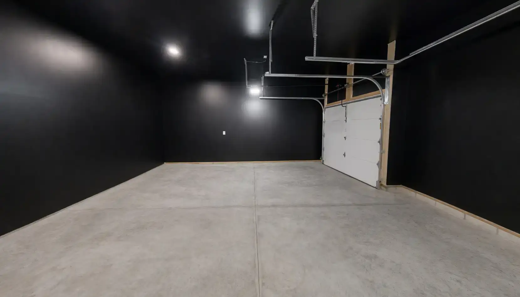 A clean, empty garage with black walls, a concrete floor, overhead lights, and a closed white garage door. The space is well-lit and spacious, featuring modern fixtures and a subtle touch of Leo-inspired boldness in its striking design—no visible objects or vehicles are present