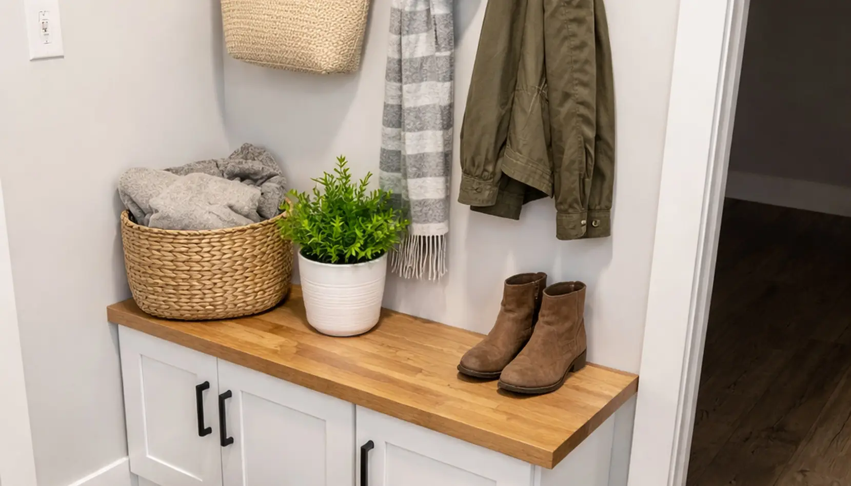 An Asher wooden bench with a white cabinet below sits invitingly, complemented by a potted plant, boots, and a woven basket with blankets. Coats and a scarf hang on hooks above, completing the cozy entryway scene