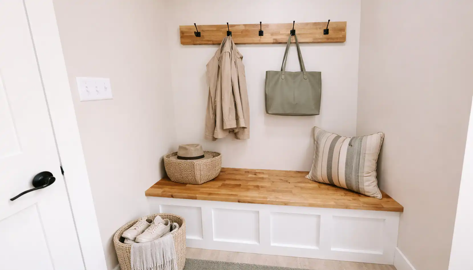 A tidy DaVinci-inspired mudroom with a wooden bench, coat hooks holding a tan coat and green bag, a striped pillow, baskets with a hat and shoes, and neutral decor