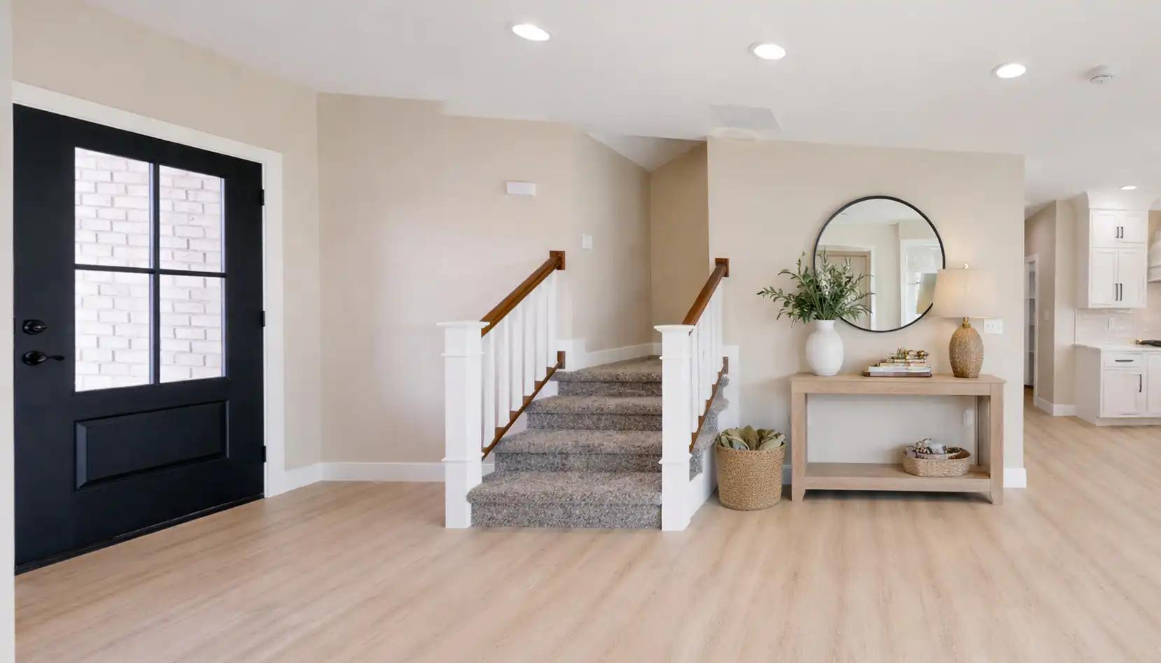 Bright entryway with a DaVinci black door, light wood floors, a carpeted staircase, a console table with decor, a round mirror above, and a basket with blankets underneath. Open kitchen visible in the background