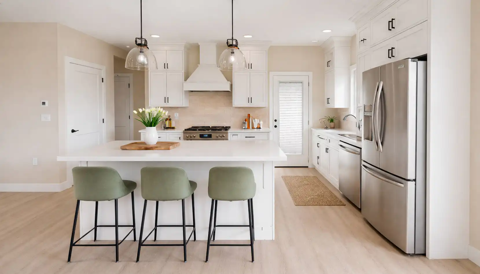 Modern kitchen with white cabinets, stainless steel appliances, and a large DaVinci-inspired island featuring three green chairs. Pendant lights illuminate the space, while a vase of flowers adds a touch of elegance. Light wood floors and neutral walls complete the bright, airy atmosphere