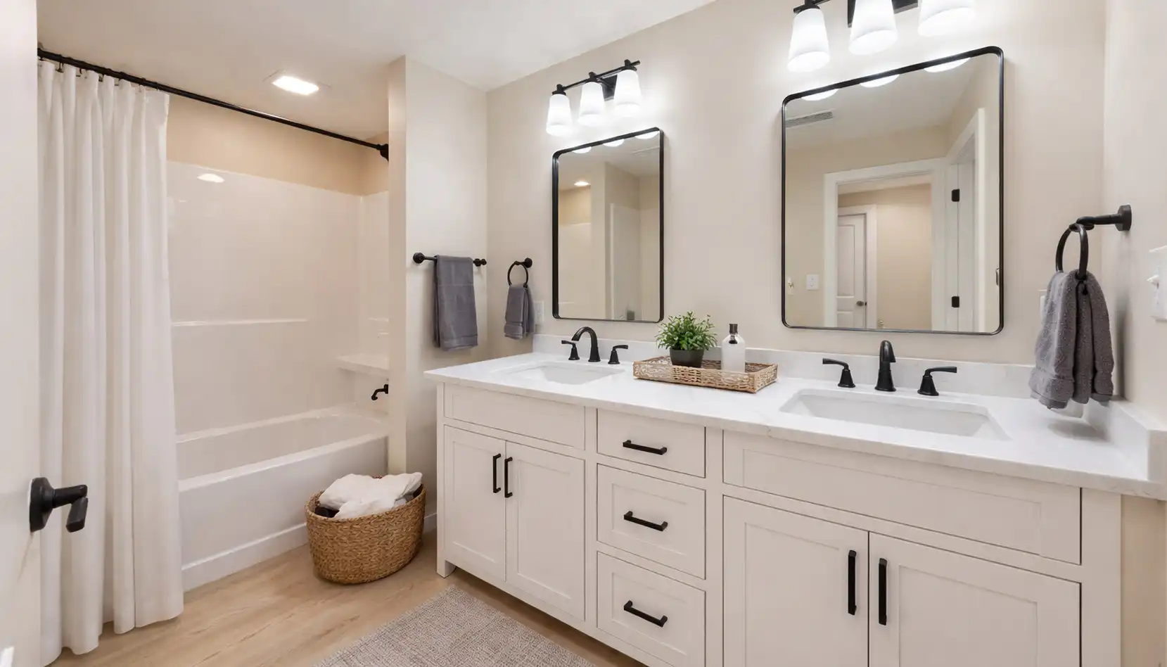 Modern bathroom with double sink vanity, white cabinets, two mirrors, black fixtures, and a DaVinci bathtub with a white shower curtain. A basket with towels sits on the floor, and a small plant decorates the countertop