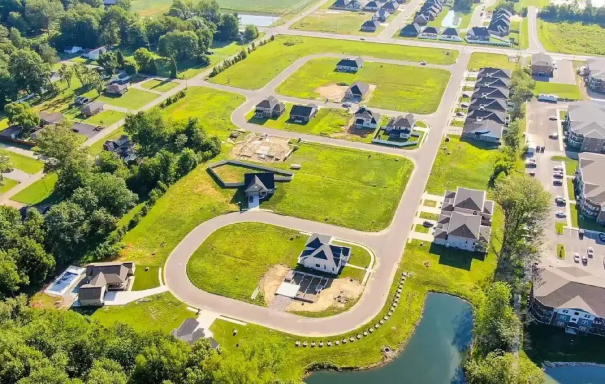 Aerial view of Pebble Creek Subdivision , a suburban neighborhood with new homes, green lawns, roads, a pond, and areas under construction. Some houses are finished, while others are partially built. Trees border the neighborhood.