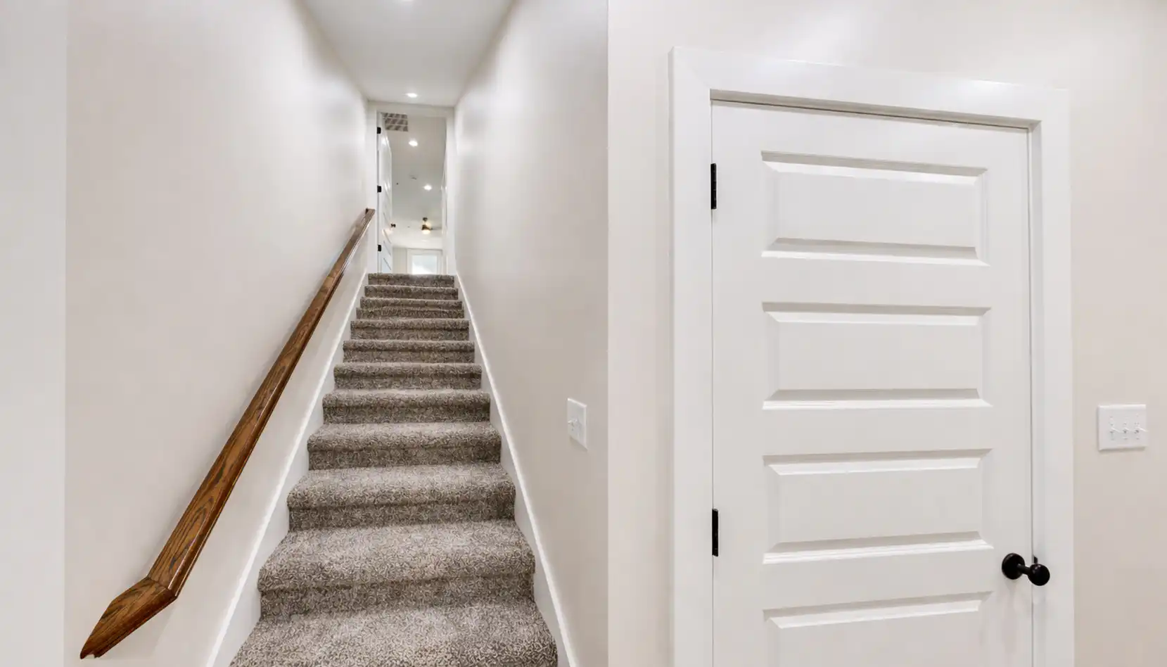 Carpeted staircase with a wooden handrail leads up a narrow hallway, reminiscent of Sophia’s timeless design sensibilities. To the right, there is a closed white door with a black handle and five horizontal panels. Walls and trim are painted white