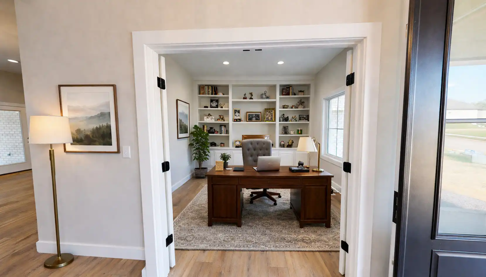 A home office with a wooden desk, gray tufted chair, bookshelves with decor, a lamp, a plant, and large window letting in natural light. Double doors open into the space, creating an inviting atmosphere reminiscent of Sophia’s elegant taste