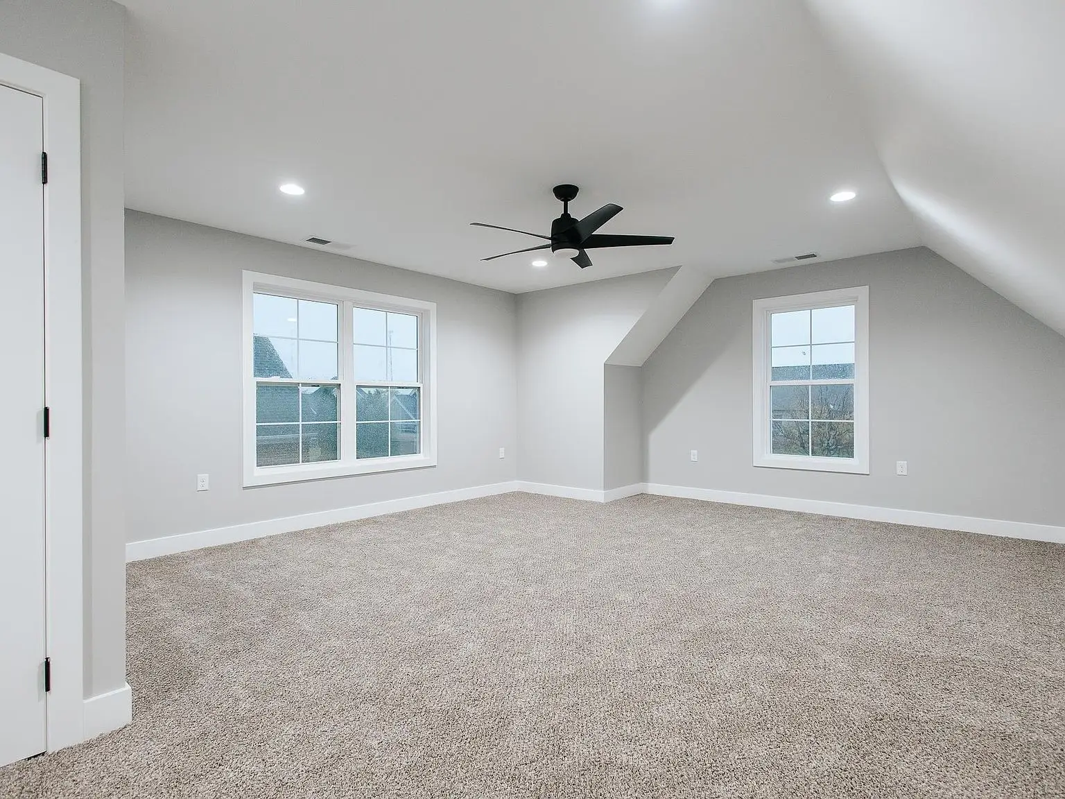 Empty carpeted room in the Eleanor floor plan with two windows, white walls, black ceiling fan.