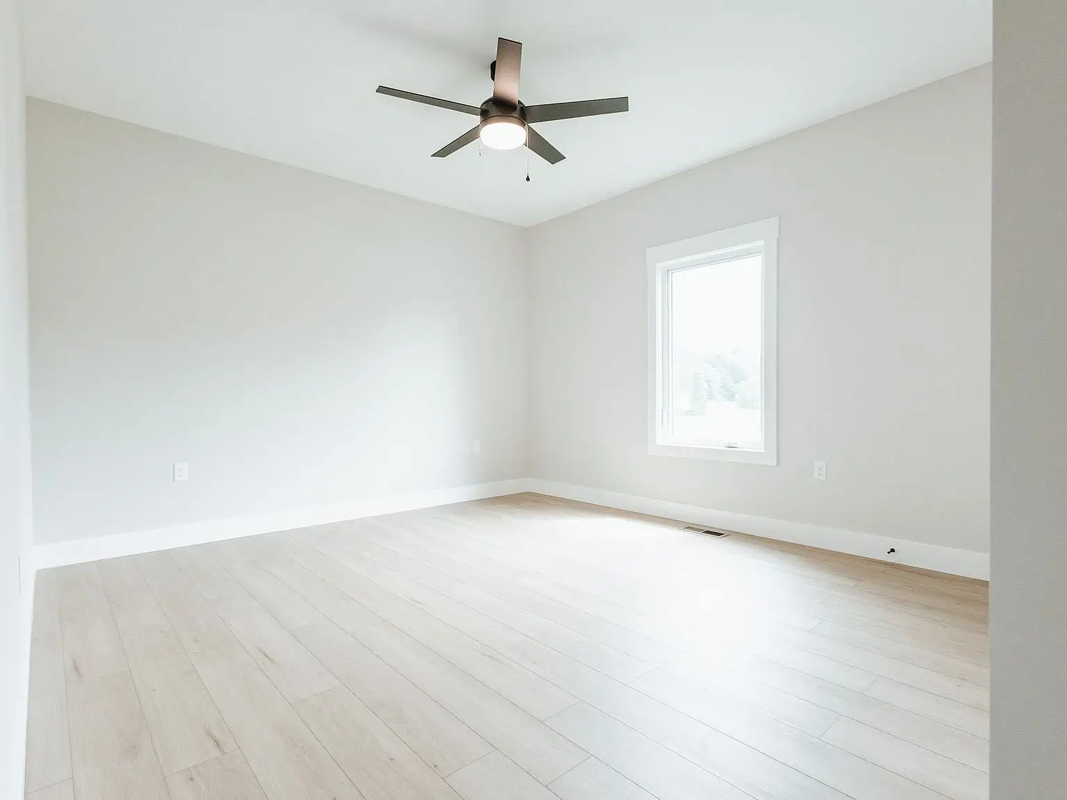 Empty room in Harper home with light wood floors, white walls, window, and ceiling fan with light.