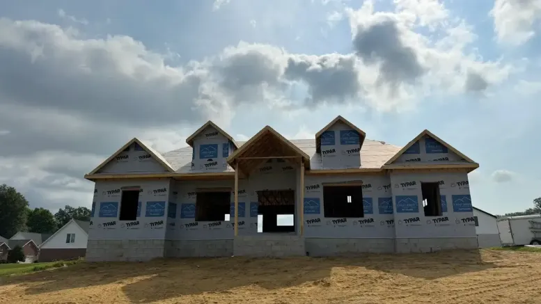 A blog-worthy scene of a house under construction with exposed framing, roof sheathing, and building wrap. The sky is partly cloudy, and the ground in front is covered with dirt.