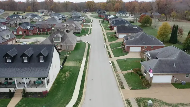 Aerial view of a suburban neighborhood with neatly lined single-family houses, vibrant green lawns, sidewalks, and a curving street, showcasing well-planned communities surrounded by autumn trees in the background.