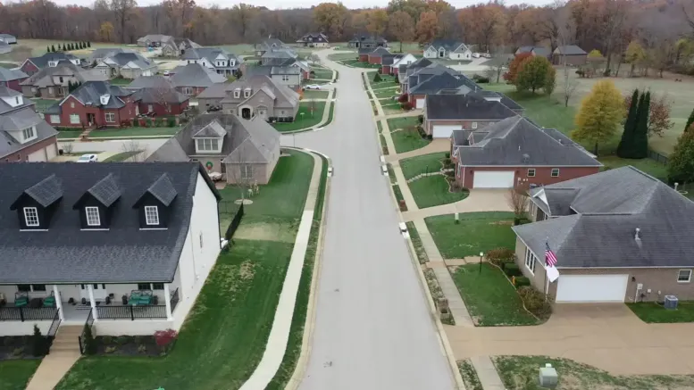 Aerial view of a suburban neighborhood with neatly lined single-family houses, vibrant green lawns, sidewalks, and a curving street, showcasing well-planned communities surrounded by autumn trees in the background.