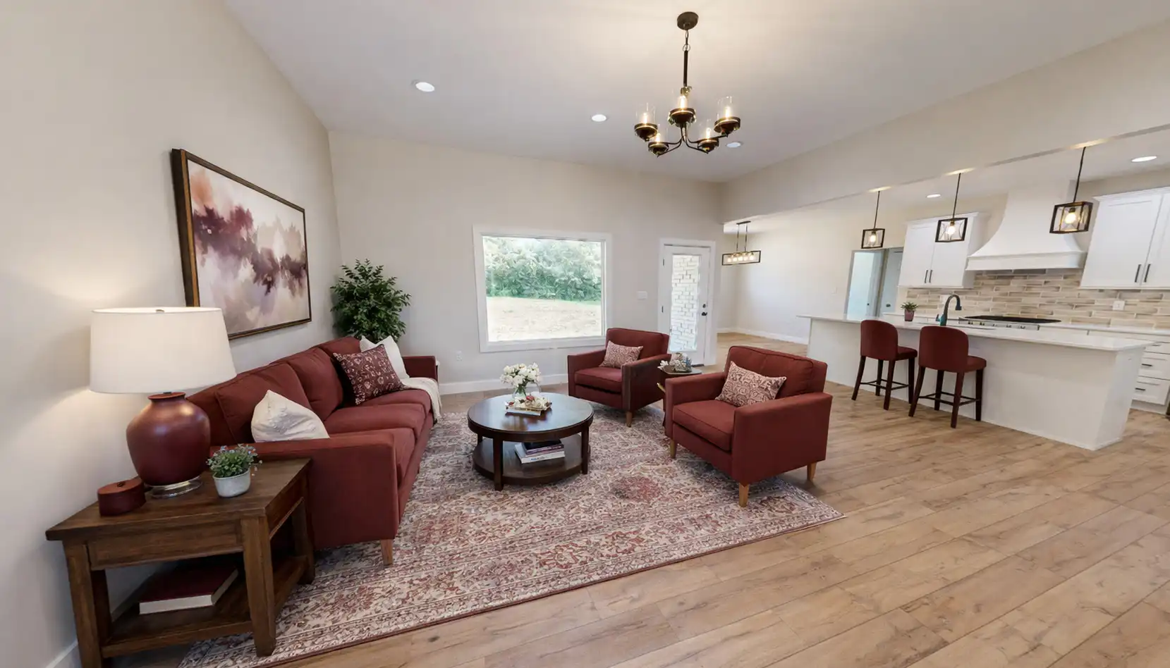 A cozy living room with red sofas, a rug, a round coffee table, and a side table with a lamp creates a welcoming Sophia-inspired ambiance. An open kitchen with bar stools and pendant lights is in the background, while large windows bring in natural light