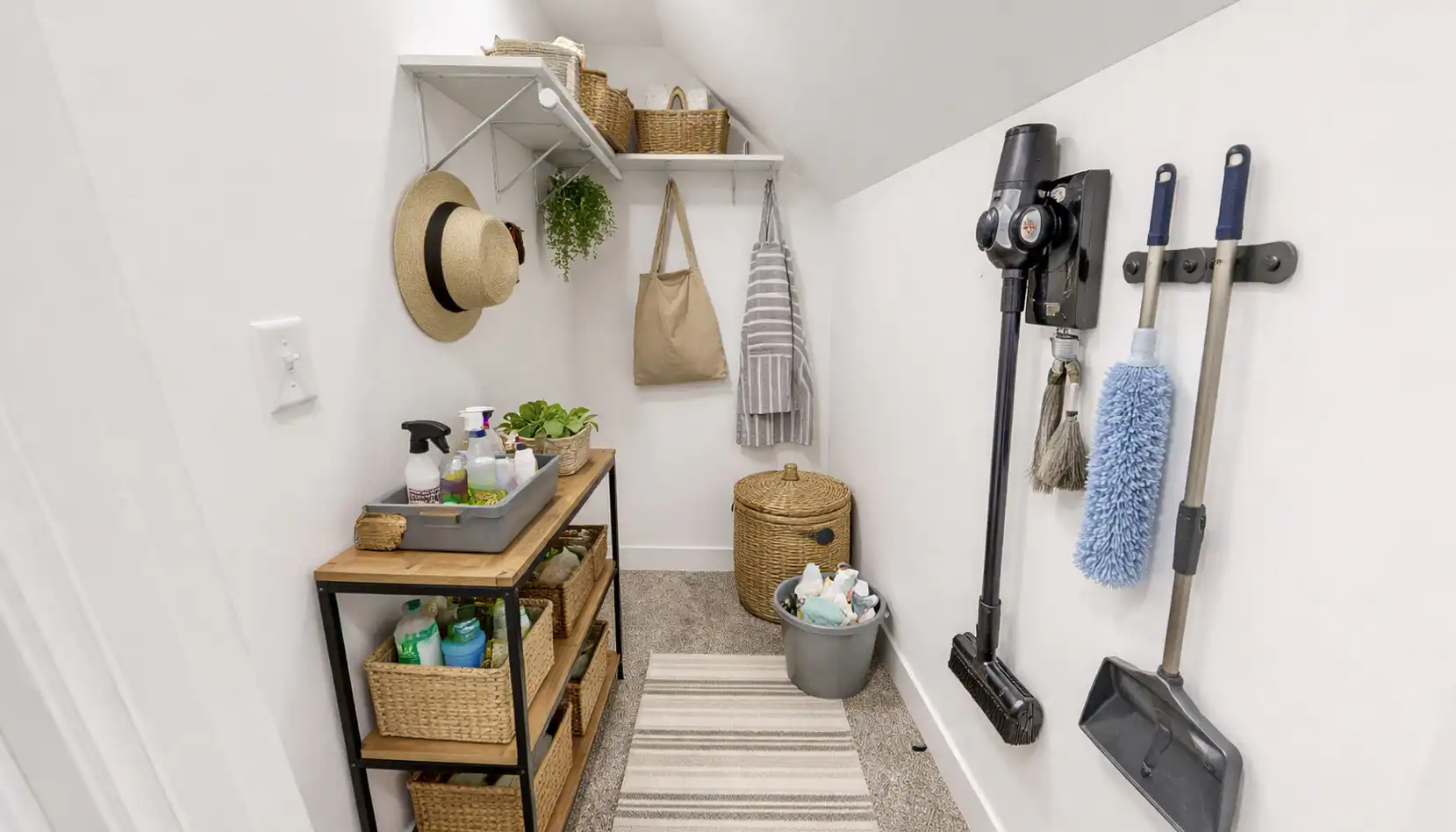 A small, organized utility room with shelves, baskets, cleaning supplies, a vacuum, hats, and a hanging bag creates an efficient space for household chores. A rug covers part of the floor while a mop, dustpan, and duster are neatly mounted on the wall—making it easy for Sophia to keep everything tidy and accessible