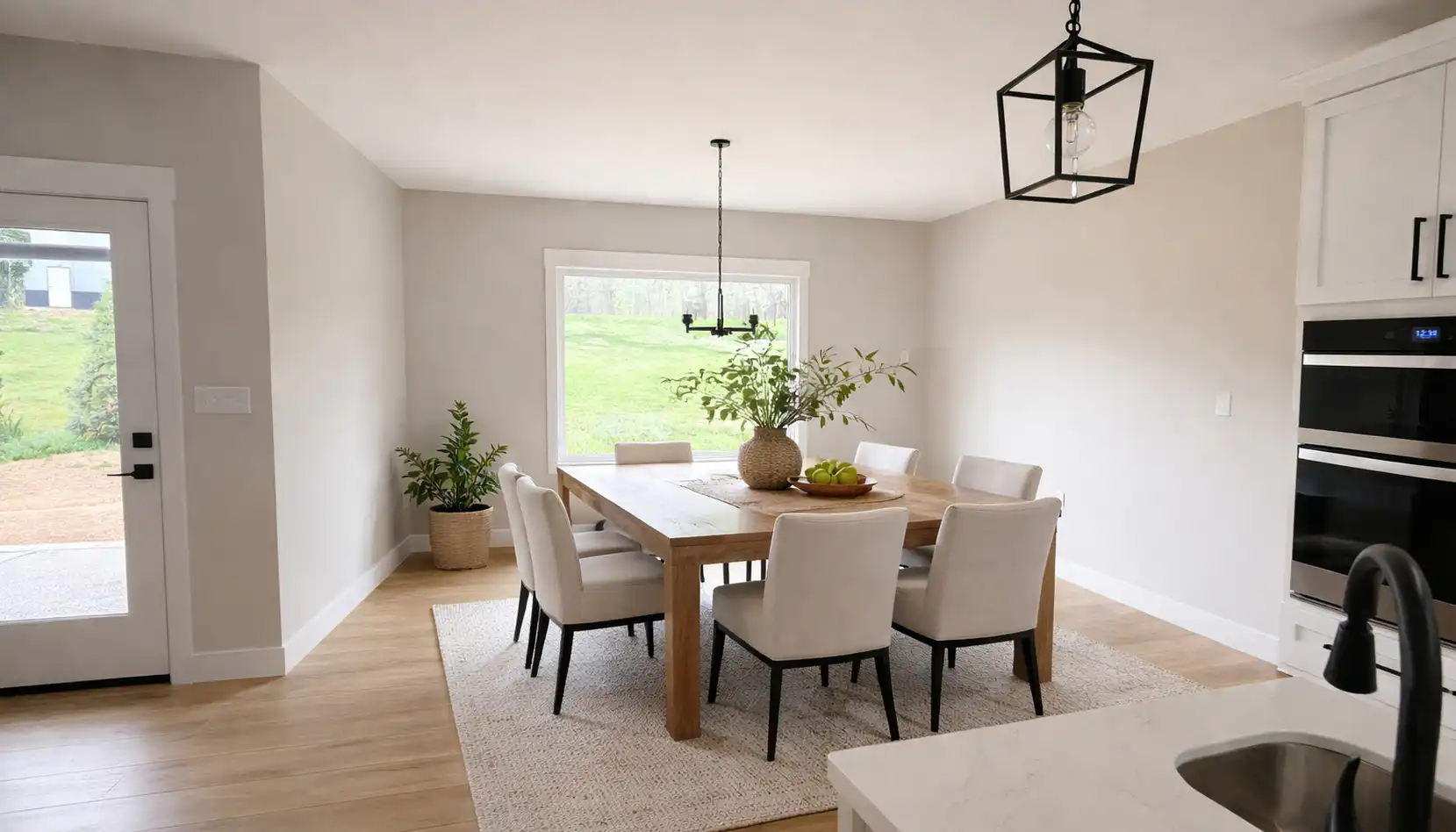Bright dining room with a wooden table, six cushioned chairs, large window, potted plants, modern light fixtures, and light wood flooring. A neutral rug sits under the table while a glimpse of robins outside the window adds a touch of nature to the inviting atmosphere