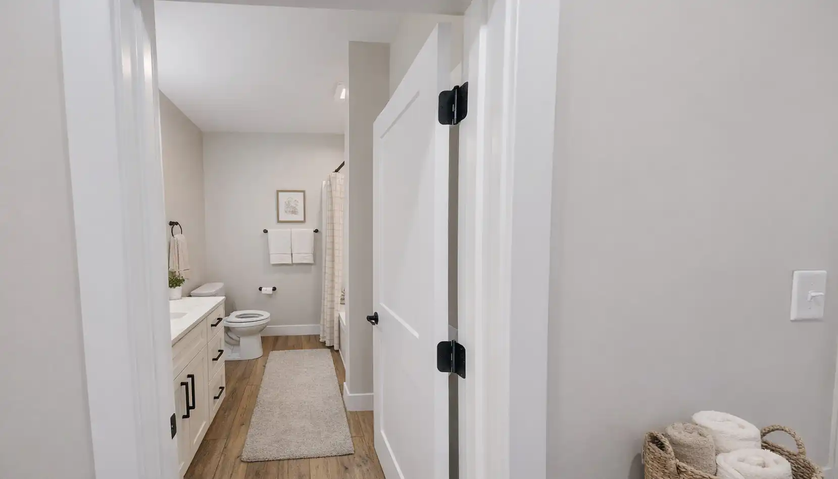 Modern bathroom with light walls, wood flooring, a white vanity, a toilet, a rug, towel racks, and a basket of rolled towels by the door. Shower curtain and framed art are visible—creating an inviting retreat that pairs perfectly with an Ava front garage home’s contemporary style
