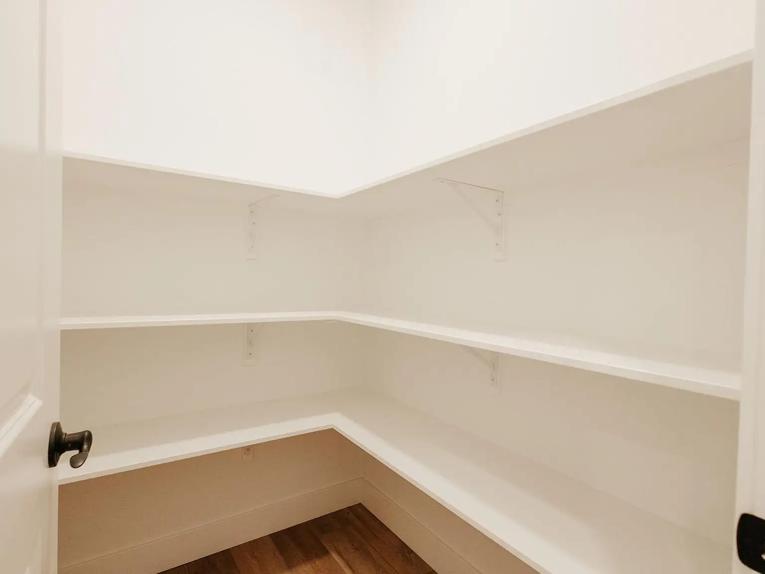 Empty white walk-in pantry with corner shelves in the Asher floor plan wood floor, open door view.