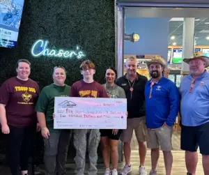 Seven people hold an oversized check, giving back to community, by a wall with a Chasers neon sign.