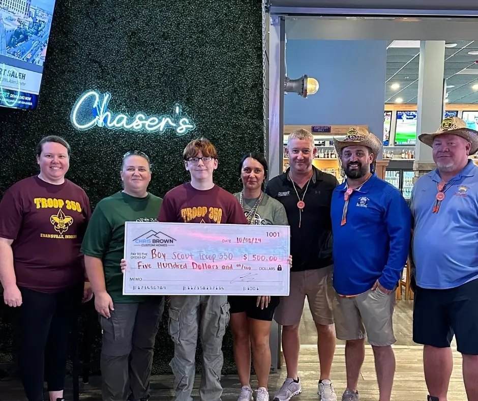 Seven people hold an oversized check, giving back to community, by a wall with a Chasers neon sign.
