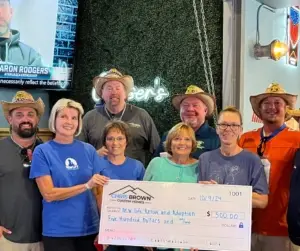 Eight people, some in cowboy hats, hold a $500 donation check, giving back to community indoors.