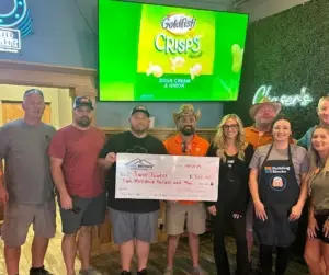 Ten people giving back to community hold a $500 check in a restaurant under a Goldfish ad.