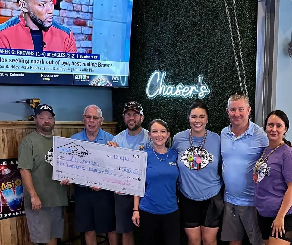 Seven people stand indoors holding a large check, giving back to community under Chasers sign.