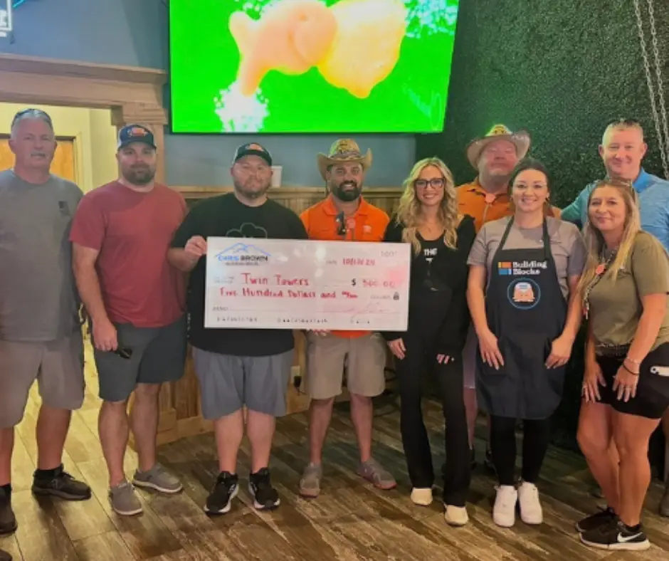 Nine people pose indoors with a large check for $502 raised for Twin Towers, restaurant setting.