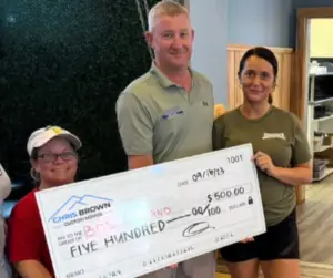 Three people hold a large check for $500 dated 09/18/23 inside a casual indoor setting.