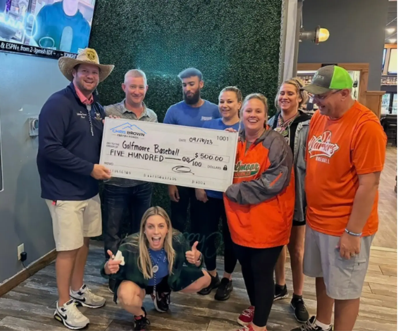 A group of people hold a $500 check for Golfmore Baseball and pose indoors, smiling.