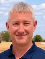 A man with short gray hair smiles outdoors on a sunny day, wearing a dark blue polo shirt. As a respected home builder in Evansville, he stands against the blurred backdrop of trees and a field under a partly cloudy sky.